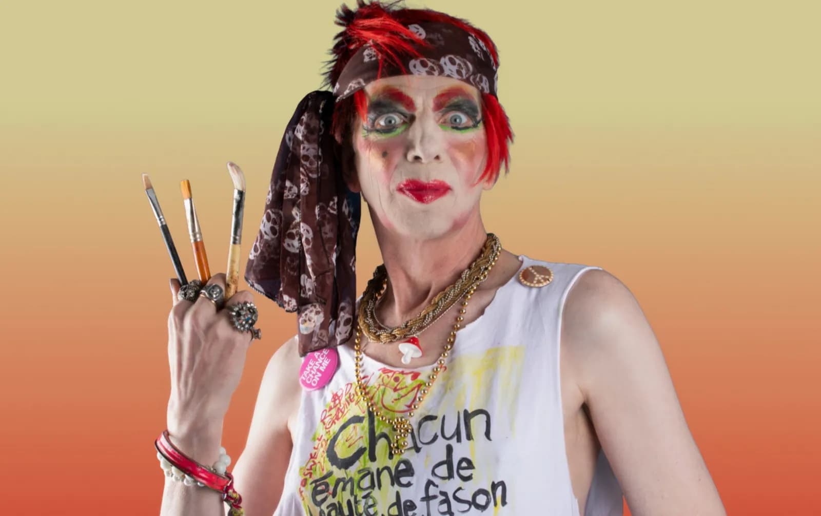A person with elaborate face paint, red spiky hair, and a bandana holds paintbrushes while wearing a white tank top with French text and several necklaces, against an orange and yellow gradient background.