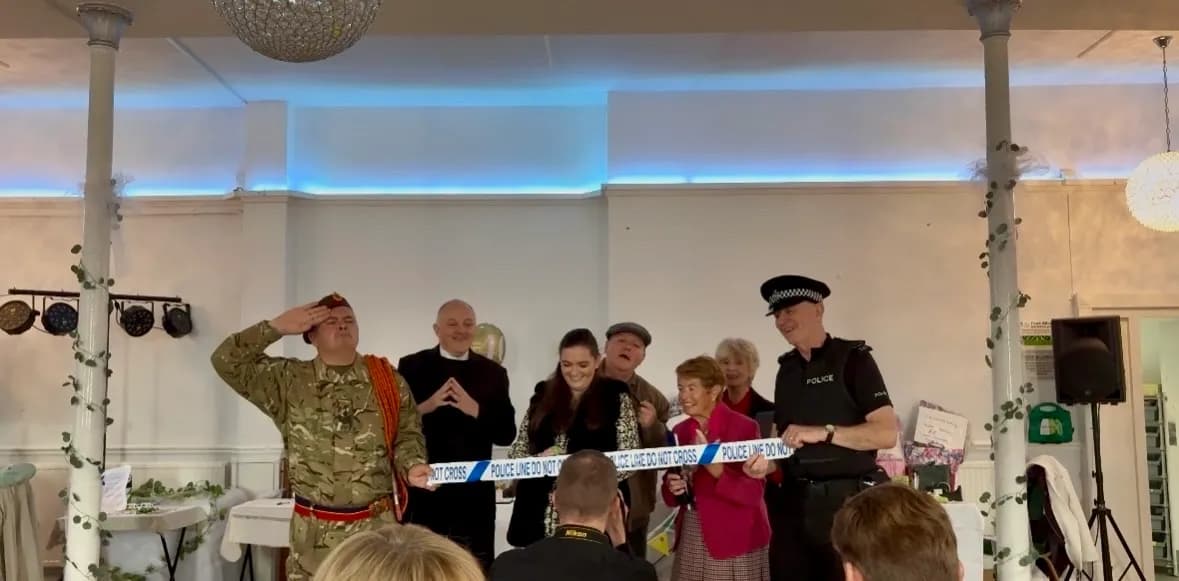 A group of people dressed in costumes, including a soldier, priest, and police officer, stand behind a blue 'POLICE LINE DO NOT CROSS' tape, smiling and engaging with an audience.