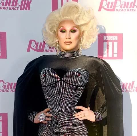 A drag queen with platinum blonde hair, dramatic eye makeup, and a black sparkling corset top, poses in front of a white and pink 'RuPaul's Drag Race' backdrop.