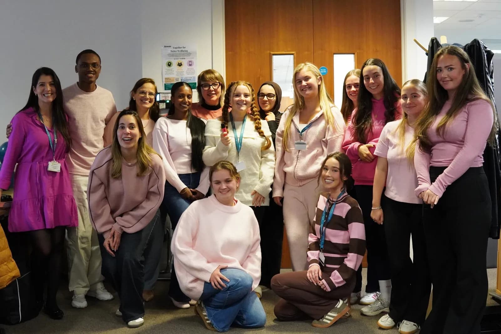 A large group of smiling young adults, mostly women, dressed in various shades of pink clothing, are gathered for a photo in an office setting.