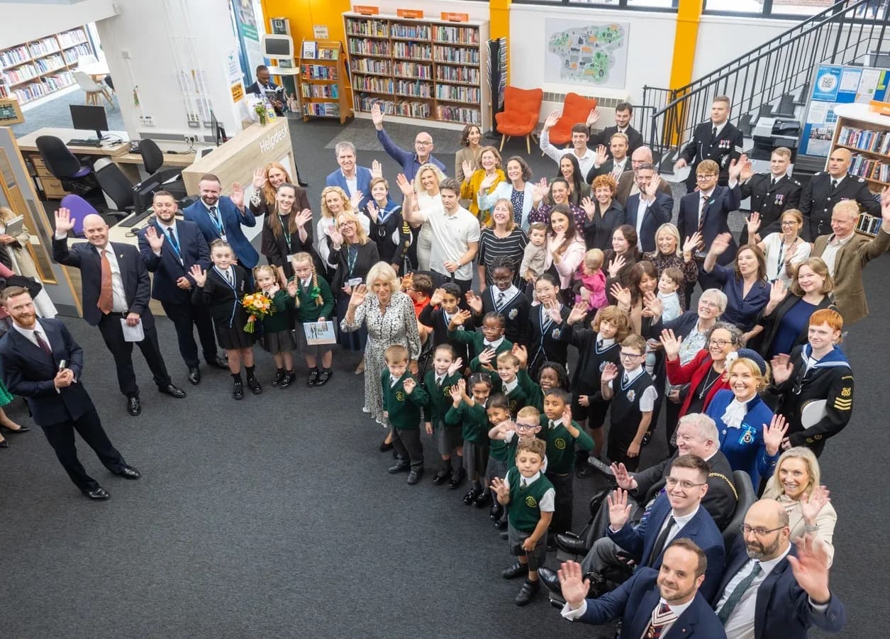 A large group of adults and uniformed schoolchildren wave at the camera in a modern library with bookshelves and desks visible in the background.